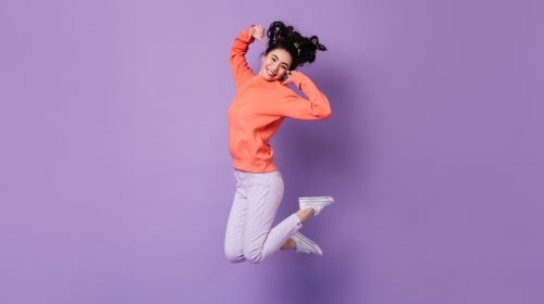 Femme souriante, sautant en l'air avec un pull orange vif, sur fond violet