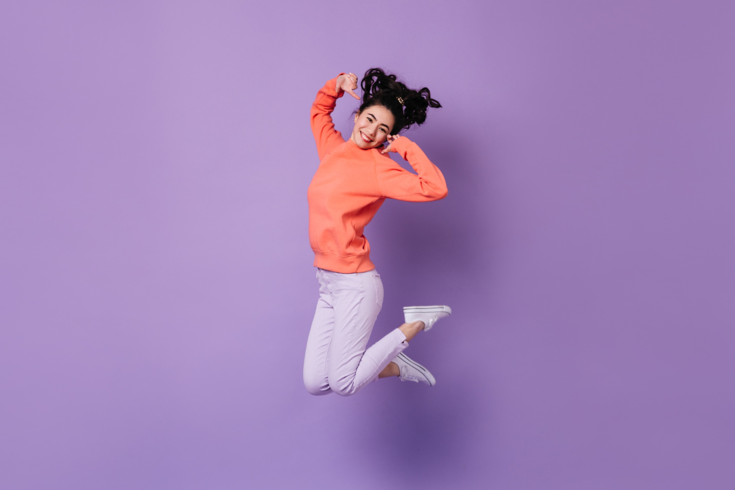 Femme souriante, sautant en l'air avec un pull orange vif, sur fond violet